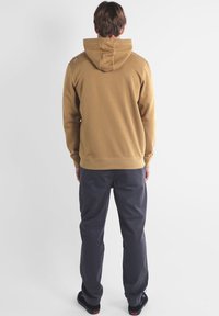 Sweatshirt à capuche marron avec une coupe décontractée, présentant un détail de couture au dos. Assorti à un pantalon gris foncé et des chaussures noires.