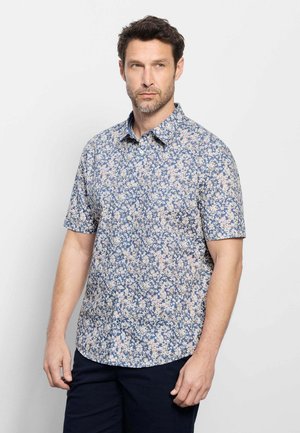 Homme portant une chemise à manches courtes boutonnée avec un motif floral bleu et blanc, debout devant un fond clair uni.