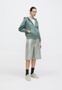 Πράσινο φούτερ με φερμουάρ και τσέπη καγκουρό, συνδυασμένο με λαμπερά ασημένια σορτς από τζιν. Το μοντέλο φοράει ανοιχτόχρωμες κάλτσες και ανοιχτόχρωμα αθλητικά παπούτσια. Μινιμαλιστικό στυλ.