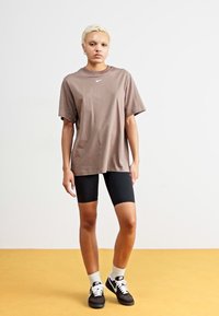 Rjava oversize majica s majhnim belim logotipom, v kombinaciji s črnimi kolesarskimi hlačami. Model nosi črne čevlje in bele nogavice. Minimalističen dizajn.