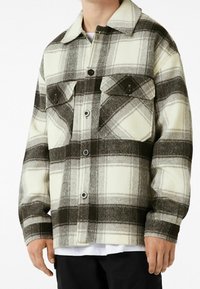 Chaqueta de cuadros en colores crema, gris y negro. Fabricada con material grueso, con dos bolsillos en el pecho y un cierre delantero abotonado. Presenta un collar clásico.