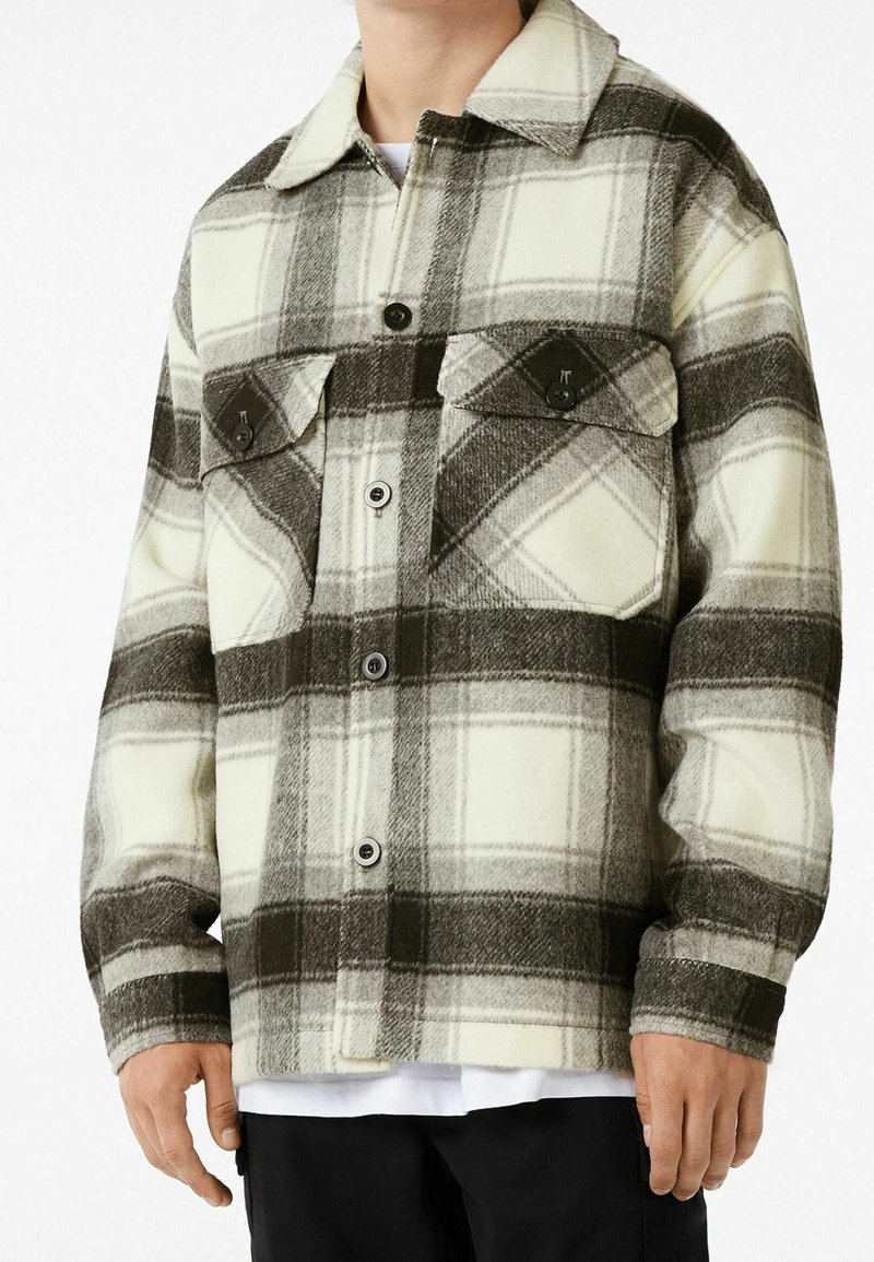 Chaqueta de cuadros en colores crema, gris y negro. Fabricada con material grueso, con dos bolsillos en el pecho y un cierre delantero abotonado. Presenta un collar clásico.