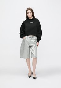 Hanorac negru scurt cu logo-ul "PINKO", combinat cu pantaloni scurți argintii lucioși până la genunchi și tocuri ascuțite negre. Modelul stă în picioare pe un fundal simplu.