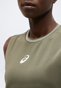 Gros plan d'une personne portant un haut de sport sans manches vert olive avec un logo Asics blanc sur la poitrine et un col rayé.