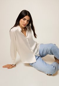Kremowa satynowa koszula z guzikami, z kołnierzykiem, długimi rękawami i luźnym krojem, w połączeniu z jasnoniebieskimi jeansami w wzory. Prosty, elegancki design.
