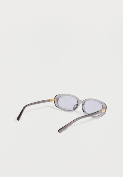 Ορθογώνια διαφανή γκρι γυαλιά ηλίου με λεπτές χρυσές λεπτομέρειες στους μεντεσέδες και τις άκρες των σκελετών, ακουμπισμένα σε φωτεινή επιφάνεια.