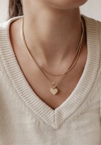Gouden gelaagde kettingen met een glad hartvormig hanger, tegen een gestructureerde crèmekleurige trui met V-hals.