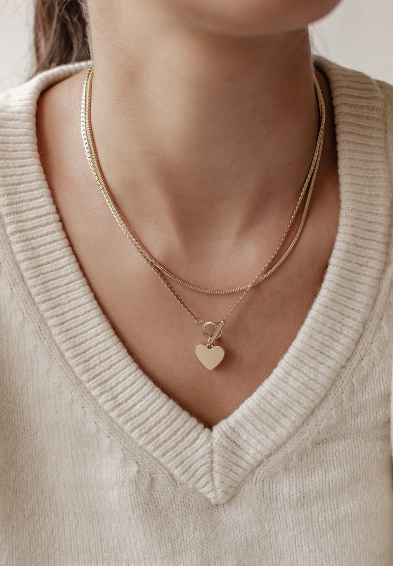 Gouden gelaagde kettingen met een glad hartvormig hanger, tegen een gestructureerde crèmekleurige trui met V-hals.