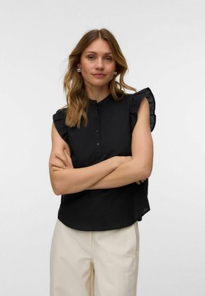 Femme aux cheveux châtain clair, portant un chemisier noir sans manches avec des volants aux épaules et un pantalon beige, bras croisés, fond neutre.