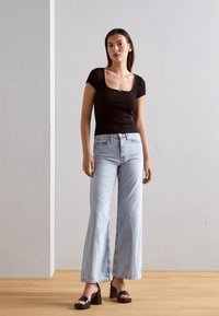Top nero a coste con maniche corte e scollatura quadrata abbinato a jeans ampi azzurri e sandali neri con plateau. Sfondo in legno.