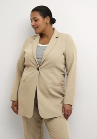 Beige přizpůsobený blejzr s jedním knoflíkem, klopami s výřezem a dvěma bočními kapsami, nošený přes pruhované tričko, s hladkou texturou tkaniny.