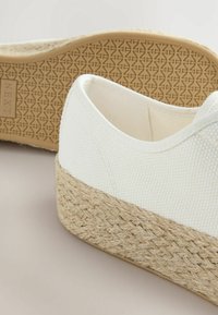 White canvas espadrille with a woven jute midsole. Features a textured rubber outsole with a geometric pattern. Minimalist design.