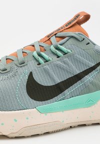 Baskets grises texturées avec une languette en mesh orange, des accents turquoise et un swoosh noir. La semelle présente un motif tacheté aux couleurs claires.