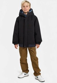 Melna ziemas jaka ar kapuci, zilas oderes un ribotas aproces, saskaņota ar brūnganām kargo biksēm un baltām sporta kurpēm ar zaļām detaļām.