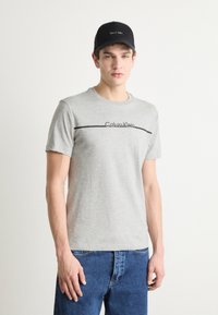 Gray cotton t-shirt with short sleeves and a black Calvin Klein logo printed horizontally across the chest. Worn with blue denim jeans.