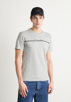 Gray cotton t-shirt with short sleeves and a black Calvin Klein logo printed horizontally across the chest. Worn with blue denim jeans.