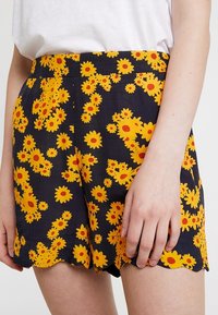 Shorts fleuris avec un fond marine orné de tournesols jaunes vifs et de cœurs rouges. L'ourlet festonné ajoute du détail au design.