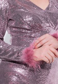 Rosa Paillettenkleid mit V-Ausschnitt, das eine strukturierte Oberfläche aufweist. Die Ärmel sind mit flauschigen rosa Pelzbündchen verziert, die das Design betonen.