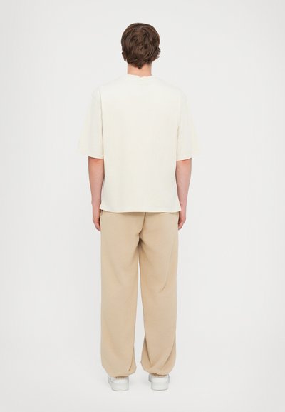 T-shirt en coton beige à manches courtes avec une coupe décontractée, associé à un pantalon de survêtement beige clair et des baskets blanches, vu de dos.