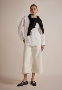 Camicia bianca con bottoni, pantaloni a gamba larga color crema, sciarpa nera e scarpe con dettagli in rete nera. Linee pulite e colori a contrasto.