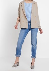 Kobieta nosząca beżową marynarkę, niebiesko-białą bluzkę w paski, dżinsy z wysokim stanem i z wywiniętymi nogawkami oraz buty na obcasie w wzór wężowy.