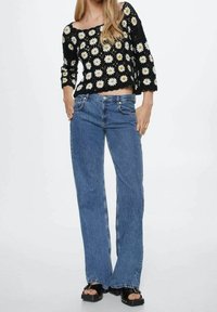 Zwarte gehaakte top met wit margrietpatroon, gecombineerd met blauwe uitlopende spijkerbroek en zwarte plateauzandalen.