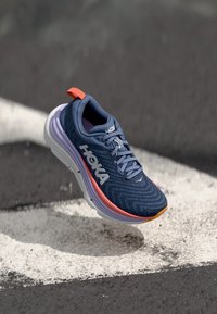 Blauer Sportschuh mit einer strukturierten Oberfläche, ausgestattet mit einer weißen und lila Sohle sowie orangefarbenen Akzenten. Enthält ein Markenlogo und ein Schnürsystem.
