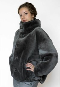 Grijze jas van nepbont met een hoge kraag, meerdere zwarte knopen en een ontspannen pasvorm. Zachte textuur en afgeronde schouders.