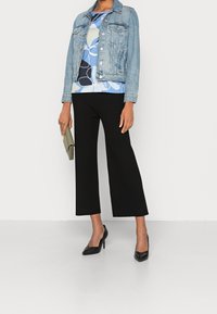 Džínová bunda v světle modré, vzorovaný top v modrých a zelených tónech, černé jemně pletené culottes a černé špičaté podpatky, držící zelenou kabelku.