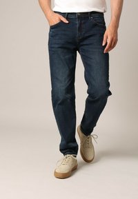 Jeans en denim bleu foncé pour hommes avec une coupe droite, des poches avant et une taille standard. Associés à des chaussures beiges à lacets.