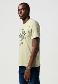 Jasnozielona bawełniana koszulka z krótkim rękawem, z czarnym nadrukiem graficznym i tekstem na przodzie. Okrągły dekolt.