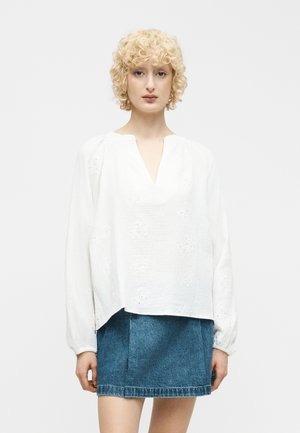 Blond kvinne med krøllete hår iført en hvit langermet bluse med hullmønster og en blå denim miniskjørt, stående mot en ensfarget bakgrunn.