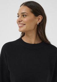 Nasmejana žena s dugom smeškom obučena u crni pulover, srebrne naušnice na obruče i tanak lančić, gleda ulijevo.