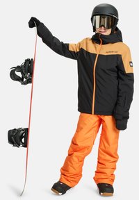 Black and orange snowboard jacket with zippers and cuffs, paired with loose orange pants. Black snowboard with black bindings carried in one hand.