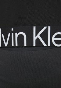 Svart åtsittande plagg med ett brett midjeband som har vit "Calvin Klein"-logotyp, tillverkat av ett mjukt och elastiskt tyg.
