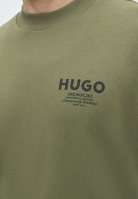 Gros plan d'une chemise vert olive avec un col côtelé et un texte noir indiquant "HUGO" ainsi qu'un lettrage stylisé plus petit en dessous sur la poitrine.