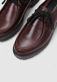 Zapatos de cuero marrón con un acabado suave, cordones negros y una suela de goma negra gruesa. Presenta costuras a lo largo de la punta y los lados.