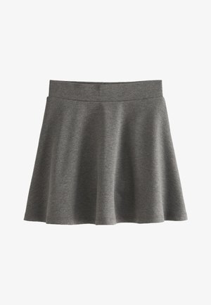 Lachend jong meisje met lang haar, gekleed in een wit shirt met korte mouwen, een grijze rok, witte sokken en zwarte schoenen met gesp, staand voor een neutrale achtergrond.
