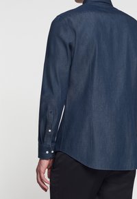 Mann trägt ein dunkelblaues Jeanshemd mit langen Ärmeln und schwarze Hose, von hinten gezeigt mit linker Hand entspannt an der Seite.