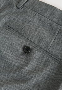 Pantalon gris sur mesure en tissu texturé, avec une poche arrière unique et un bouton noir à quatre trous en guise d'accent.