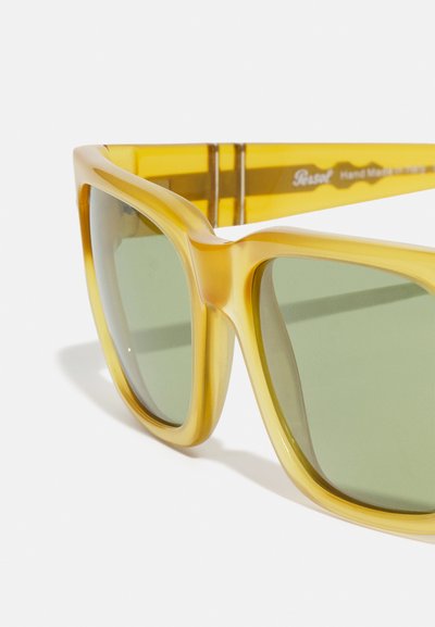 Occhiali da sole di colore giallo con montatura spessa e lenti verdi. Presentano una superficie liscia e un design angolare, con un branding discreto sulla stecca.