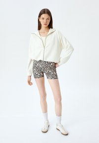 Witte zip-up cropped jas, leopard print strakke shorts, witte sneakers met bruine zolen en witte sokken. Glad materiaal, casual stijl.