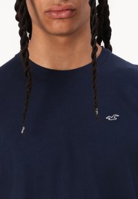 Marineblaue Baumwoll-T-Shirt mit klassischem Rundhalsausschnitt, ausgestattet mit einem kleinen, weißen Logo, das auf der linken Brust gestickt ist, und kurzen Ärmeln.