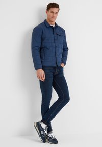 Navy quilted jacket with a collared neckline, front buttons, single chest pocket, paired with dark denim jeans and navy sneakers.
