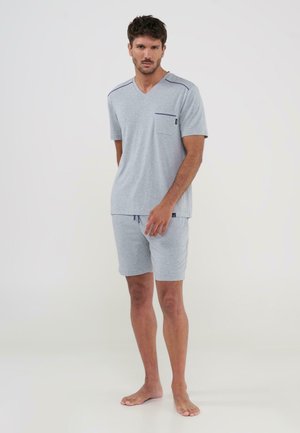 Hombre de pie descalzo con camiseta de manga corta y cuello en V color gris claro con bolsillo en el pecho y pantalones cortos a juego con cordón, sobre un fondo liso.