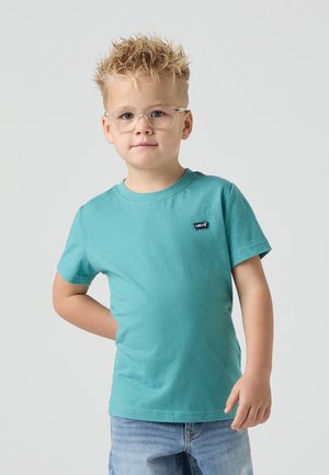 Teal bavlnené tričko s okrúhlym výstrihom, krátkymi rukávmi a malým čiernym logom na hrudi. Model stojí s jednou rukou na boku v modrých džínsových šortkách.