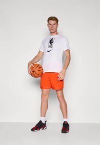 Λευκή κοντομάνικη μπλούζα με λογότυπο NBA, πορτοκαλί σορτς και μαύρα-κόκκινα αθλητικά παπούτσια. Το άτομο κρατάει μια μπάλα μπάσκετ, στέκεται μπροστά σε απλό φόντο.