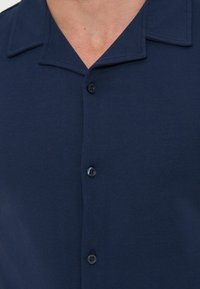 Μπλε ναυτικό πουκάμισο με κουμπιά και γιακά κάμπινγκ που φοριέται από άτομο, εμφανίζοντας το πάνω μέρος του στήθους και το λαιμό.