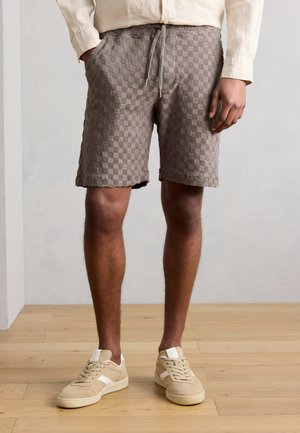 Mann iført taupe ruterte, teksturerte shorts med snøring foran, beige langermet skjorte og beige joggesko, stående på tregulv.