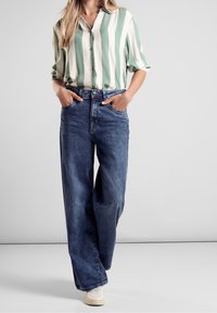 Camisa de botones a rayas verdes y blancas, combinada con unos vaqueros de mezclilla azul holgados y zapatillas blancas, diseñada para un ajuste relajado.